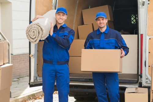 Two professional movers from Hampstead Man with Van, dressed in blue uniforms and matching caps, standing outside a residential property with an open removals van behind them. The mover on the left is holding a rolled-up carpet with a textured beige fabric, smiling at the camera, while the mover on the right is carrying a cardboard box. The van's interior is visible, filled with stacked cardboard boxes of various sizes and some furniture pieces, indicating an ongoing house removal process in a Hampstead or nearby NW postcode area. The scene is set on a residential street, with a driveway and a brick wall partly visible, and natural daylight illuminating the scene. The image emphasizes careful handling of household belongings such as soft furnishings and boxes, aligned with expert house removal services in the Hampstead area.
