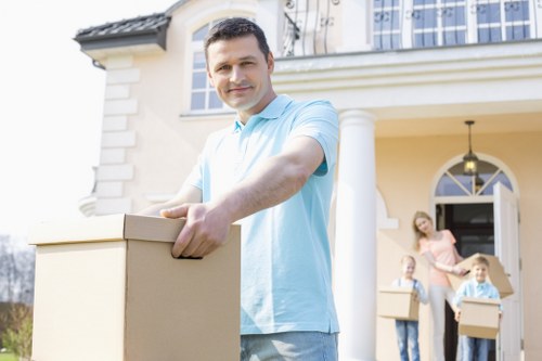 A man with short dark hair, dressed in a light blue shirt, stands outdoors in front of a modern, light-colored house with white columns and a large front door. He is smiling and holding a cardboard box with both hands, facing the camera. In the background, a woman and a child, both casually dressed, are visible carrying additional cardboard boxes towards the house, suggesting a moving or house removal activity. The scene is set on a bright day, with natural light illuminating the house façade and the individuals, emphasizing a professional but approachable environment for residential removal services in Hampstead. The house's exterior features a neatly maintained facade with arched windows and decorative architectural details, with the scene capturing the moment of packing or unpacking during a house move that Hampstead Man with Van might facilitate.