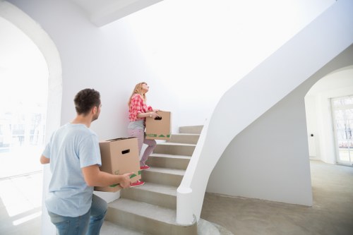 A young man and woman are engaged in a house move inside a bright, modern home with white walls and large windows. The man, dressed in a light blue t-shirt, is standing on the ground floor holding a medium-sized cardboard box, while the woman, wearing a pink checkered shirt and jeans, is ascending a curved staircase with white balustrades, carrying a similar cardboard box. Natural light floods the space through the windows, creating an airy atmosphere. The staircase features concrete steps and a smooth, white handrail, and the background shows an open, minimalist interior with clean lines and a concrete floor. These activities are part of a professional house removal, suggesting a relocation service likely offered by Hampstead Man with Van in the Hampstead area, with the setting indicating a residential property in London’s Hampstead postcode district.