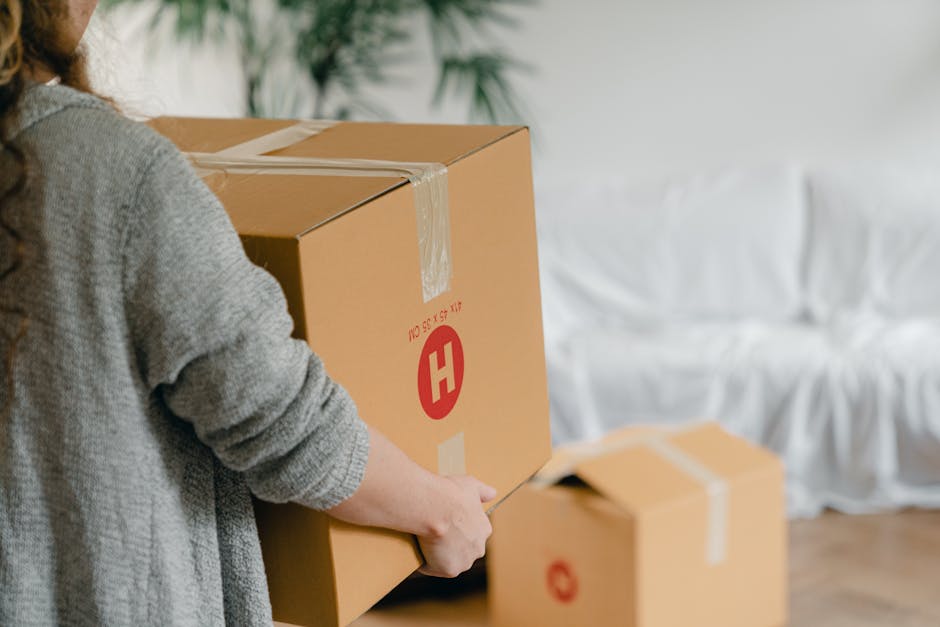 A person wearing a grey sweater is in the process of packing or unpacking moving boxes inside a residential living room. They are holding a medium-sized cardboard box sealed with clear packing tape, w
