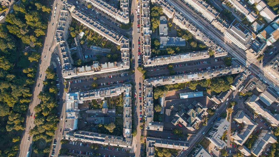 An aerial view of a densely built urban area showing rows of terraced houses along narrow streets. Narrow pavements are visible alongside the buildings, with some parked cars lining the roads and a fe